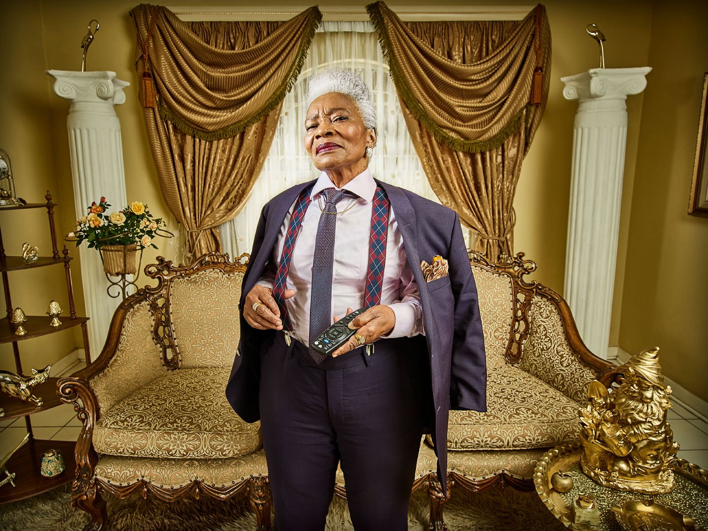 an elderly woman with boss attitude dressed in a tailored suit in a luxurious living room holding the tv remote