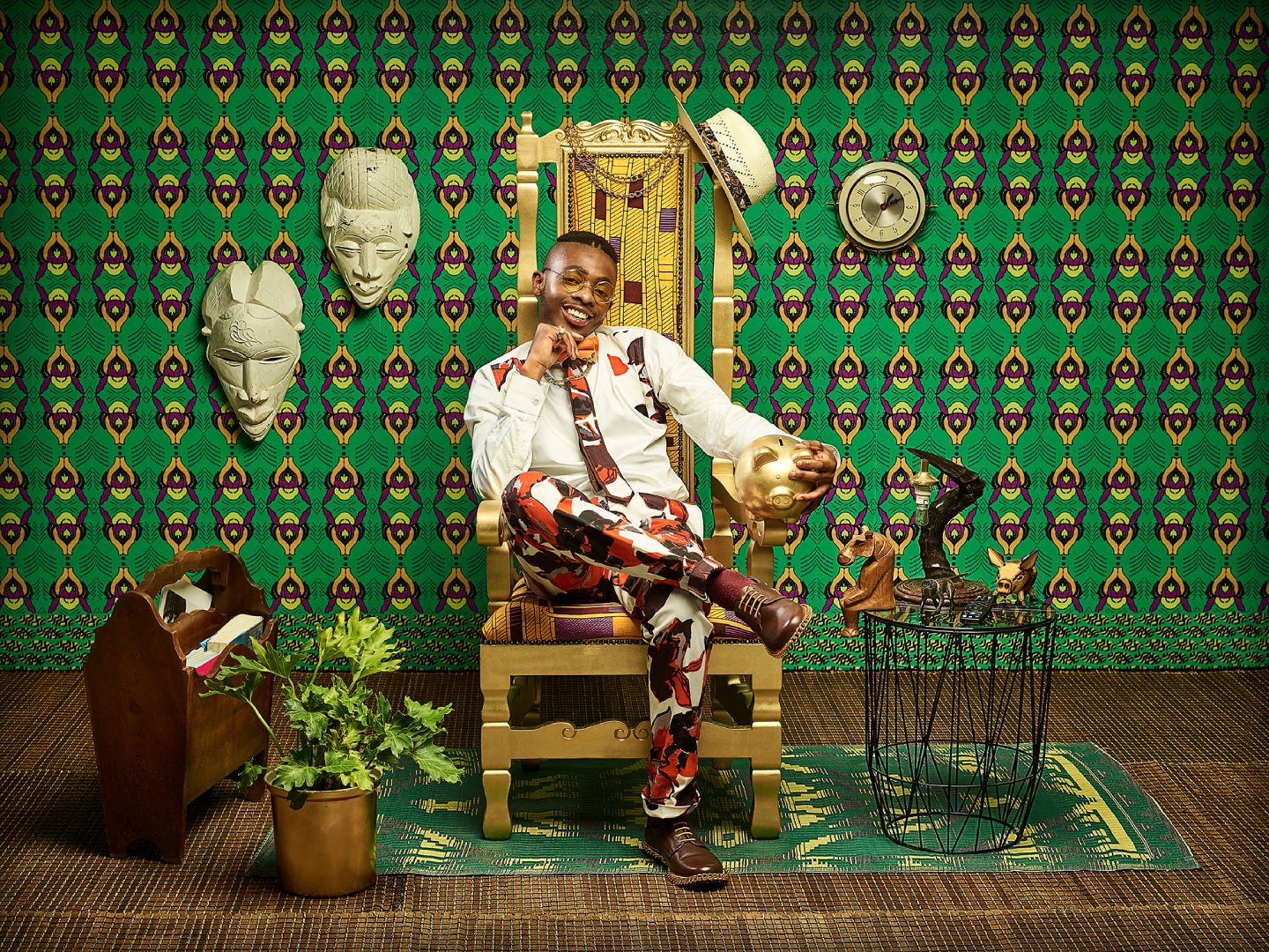 A young African man seated on a golden chair in front of a bright green African patterned wall with white masks