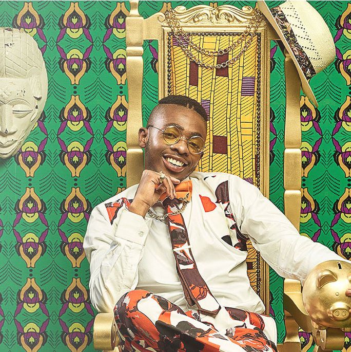 A young African man seated on a golden chair in front of a bright green African patterned wall with white masks